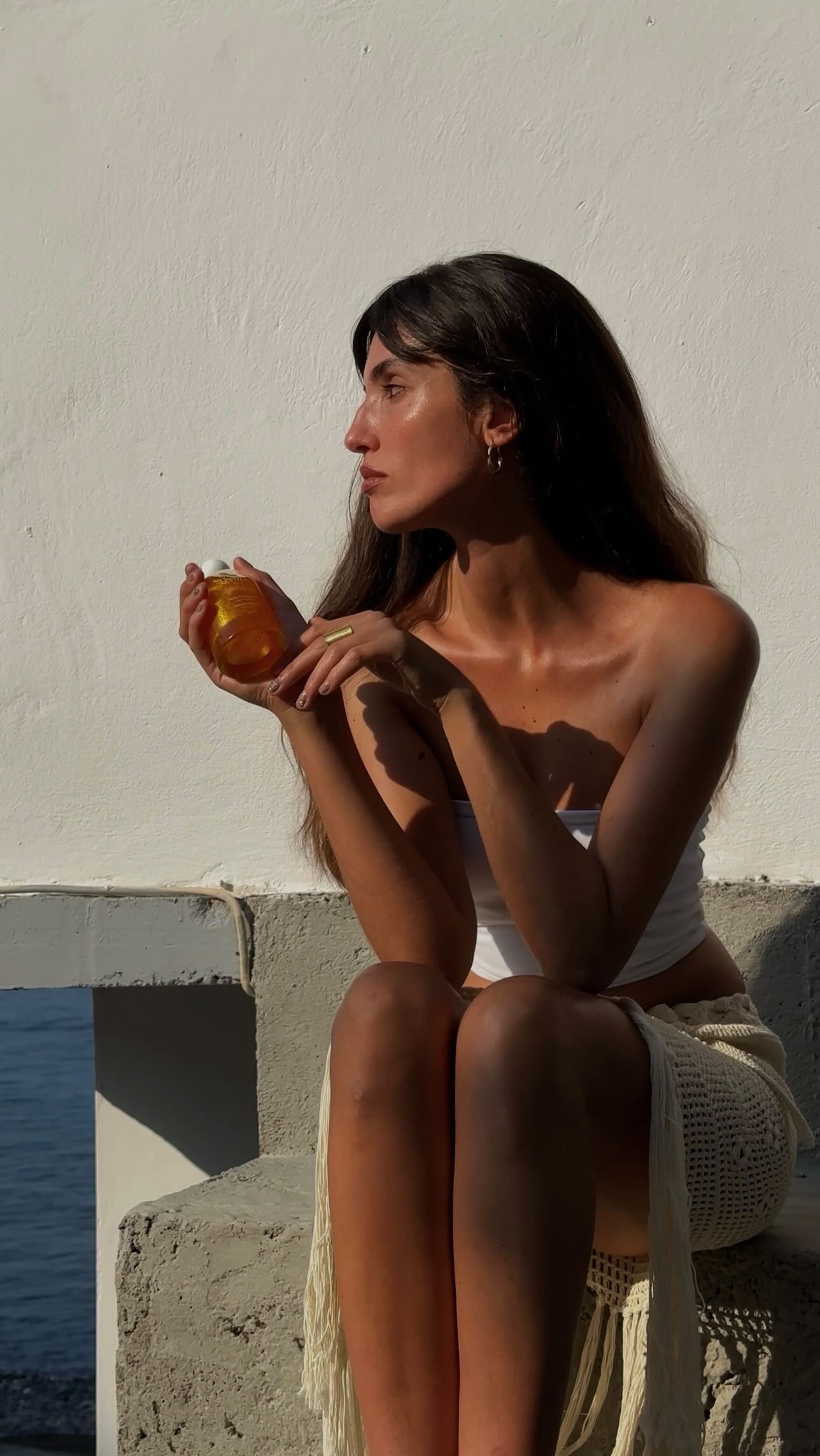 Femme assise en hauteur à proximité de la mer tenant dans sa main un produit cosmétique. 