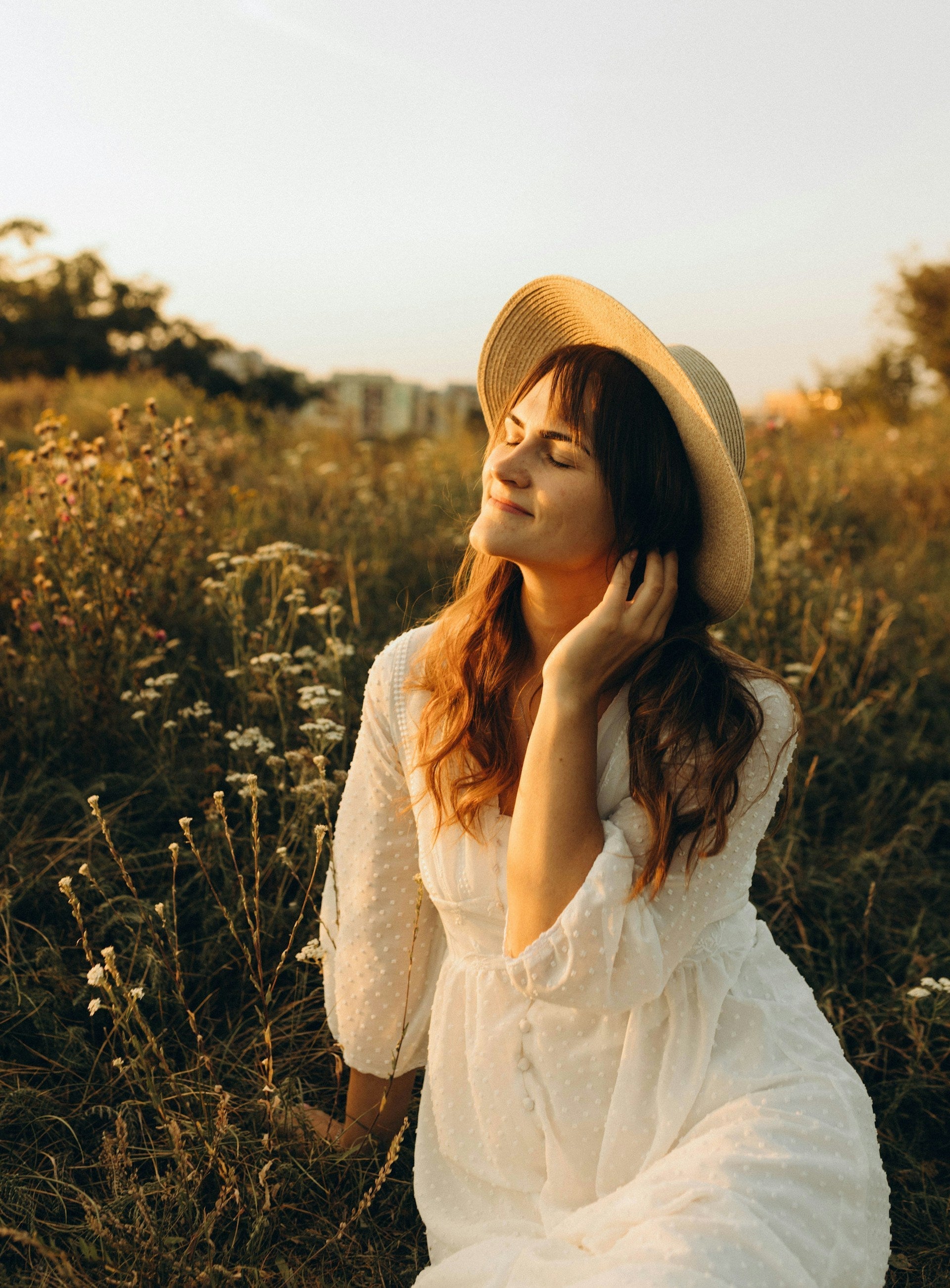 Femme en robe chemisier blanc et chapeau sur la tête assise dans un champ fleuri. 