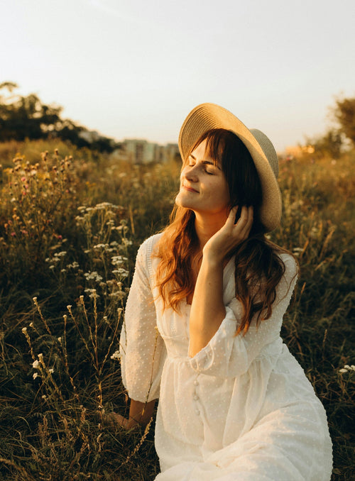 Femme en robe chemisier blanc et chapeau sur la tête assise dans un champ fleuri. 