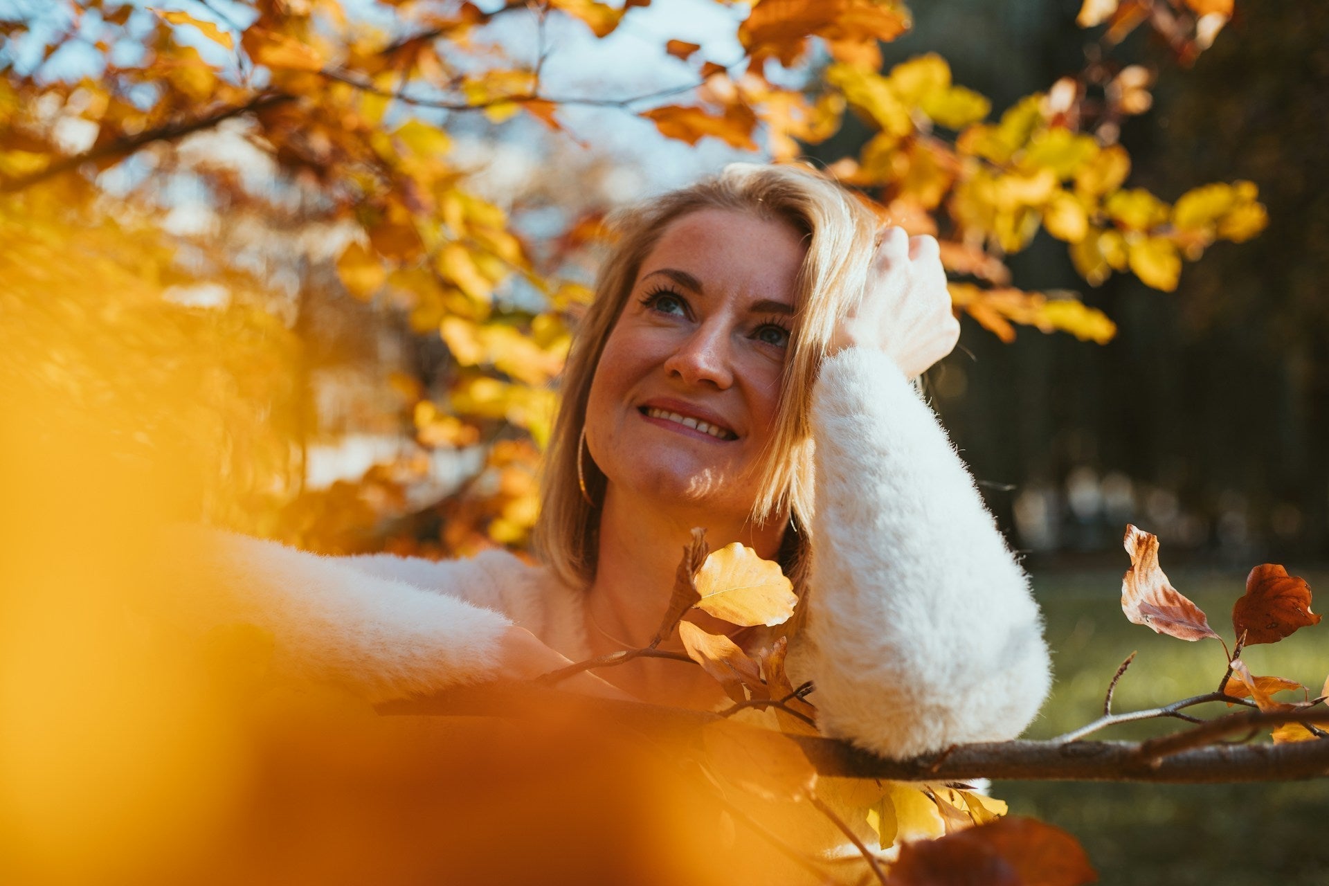 Une femme souriante en pull blanc dont le coude est posé sur une branche d’arbre.